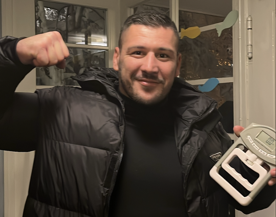 Smiling man flexing one arm while holding a digital grip strength dynamometer