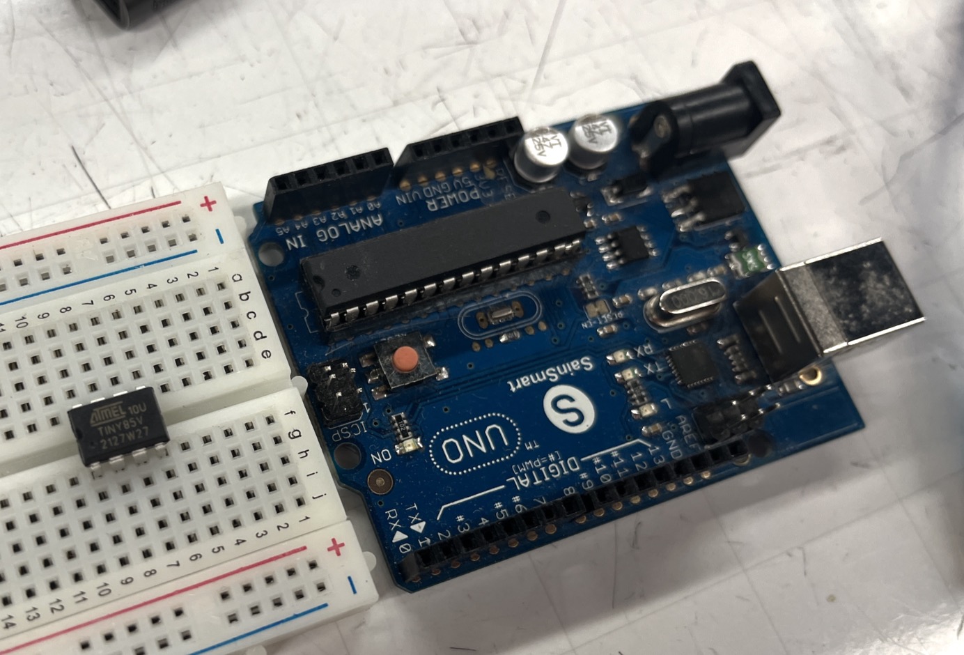 ATtiny85V chip on a breadboard next to a blue Arduino Uno board
