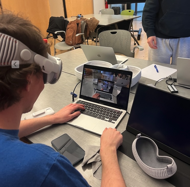 Developer typing on a MacBook while wearing the Vision Pro, laptop screen showing the headset passthrough view