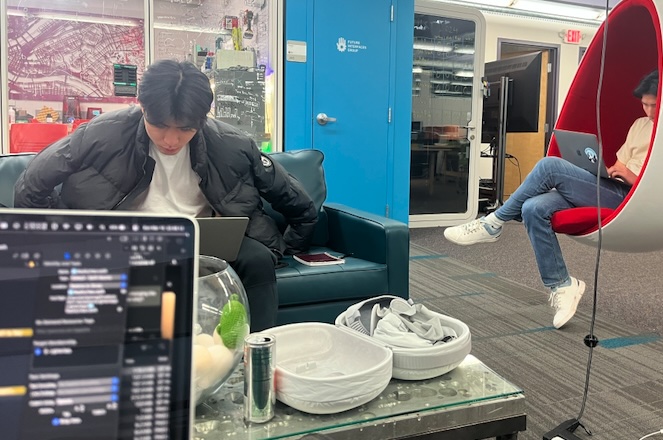 Two hackers working on laptops, one on a couch and one in a red egg chair, Vision Pro cases on the table