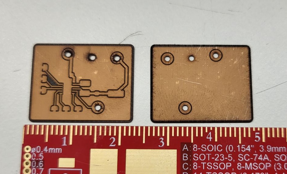 Front and back of a small milled copper breakout board for the R1801K, laid on a ruler for scale