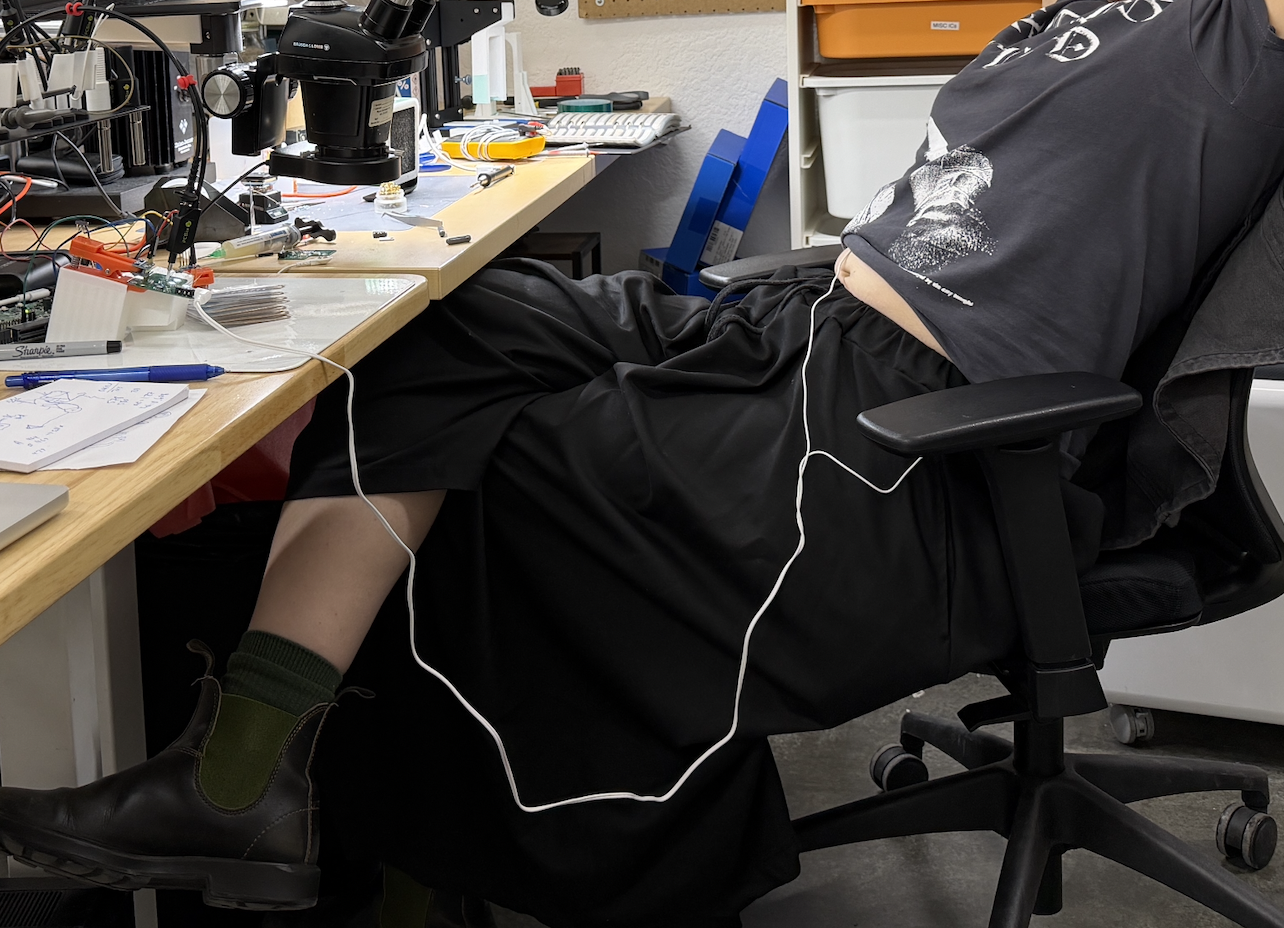 Person reclined at an electronics workbench with a long white electrode wire trailing from under their shirt to the desk