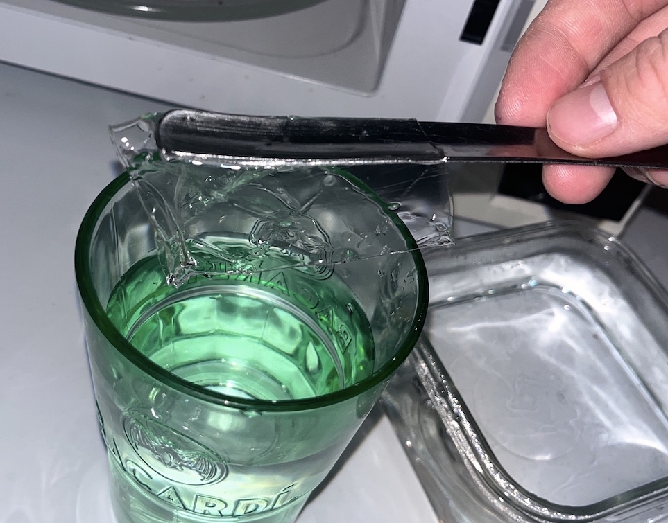Lifting the floppy soaked gelatin sheet out of a green glass of water with a knife, ready to transfer it