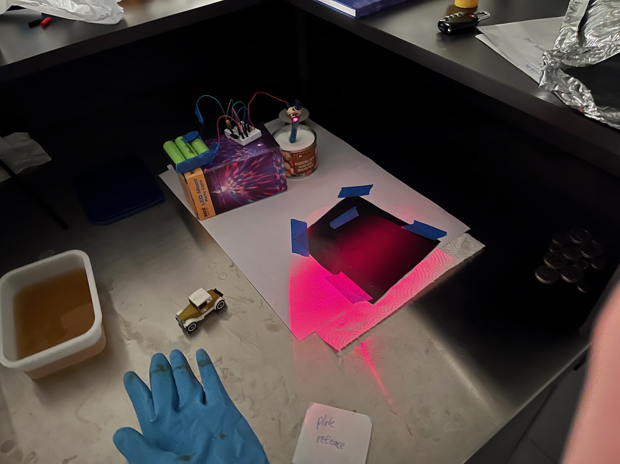 Darkened bench holography setup with a red laser spread across a taped-down black plate, toy car and rubber glove nearby