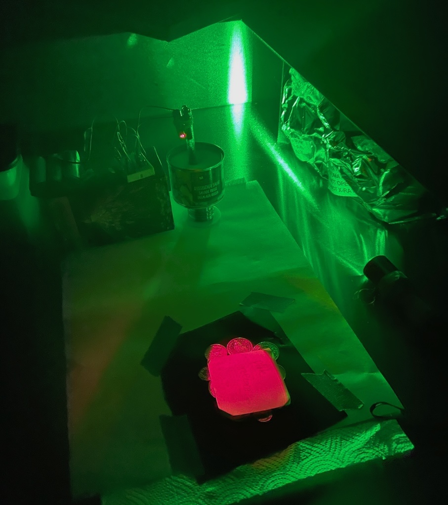 Hologram plate glowing pink over a pile of coins during laser exposure, the room lit only by a green safelight