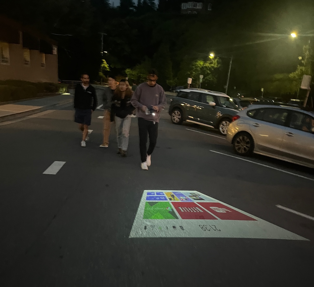 People walking at night beside a mini-projector casting an app grid onto the street pavement