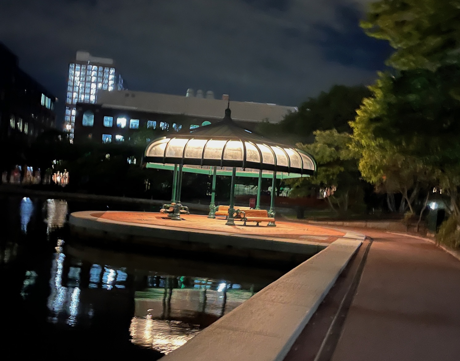Glass-roofed pavilion glowing at night beside a canal walkway, city buildings lit behind