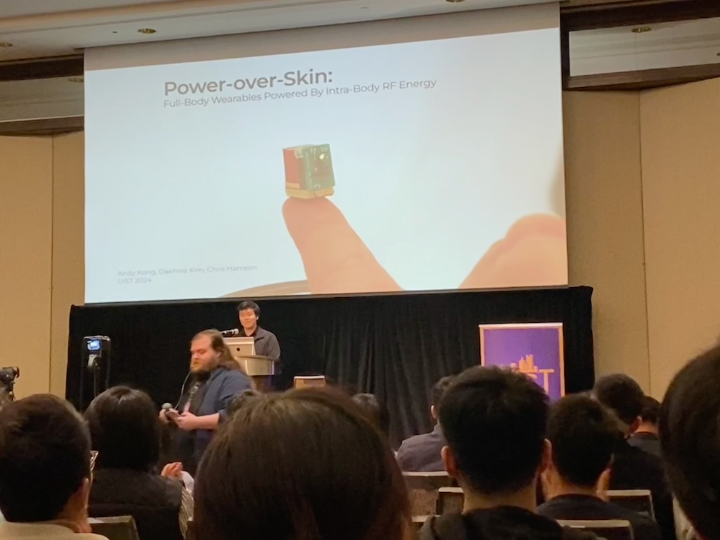 Andy at the UIST podium beneath a Power-over-Skin title slide showing a tiny device on a fingertip