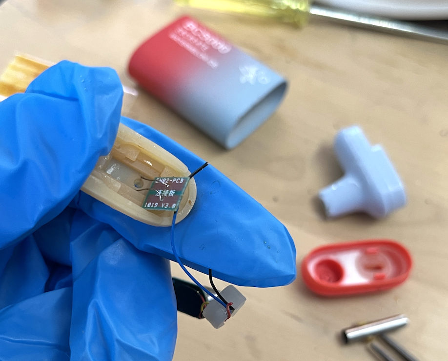 Gloved hand holding a disassembled vape pen's tiny PCB, with the casing and mouthpiece scattered behind